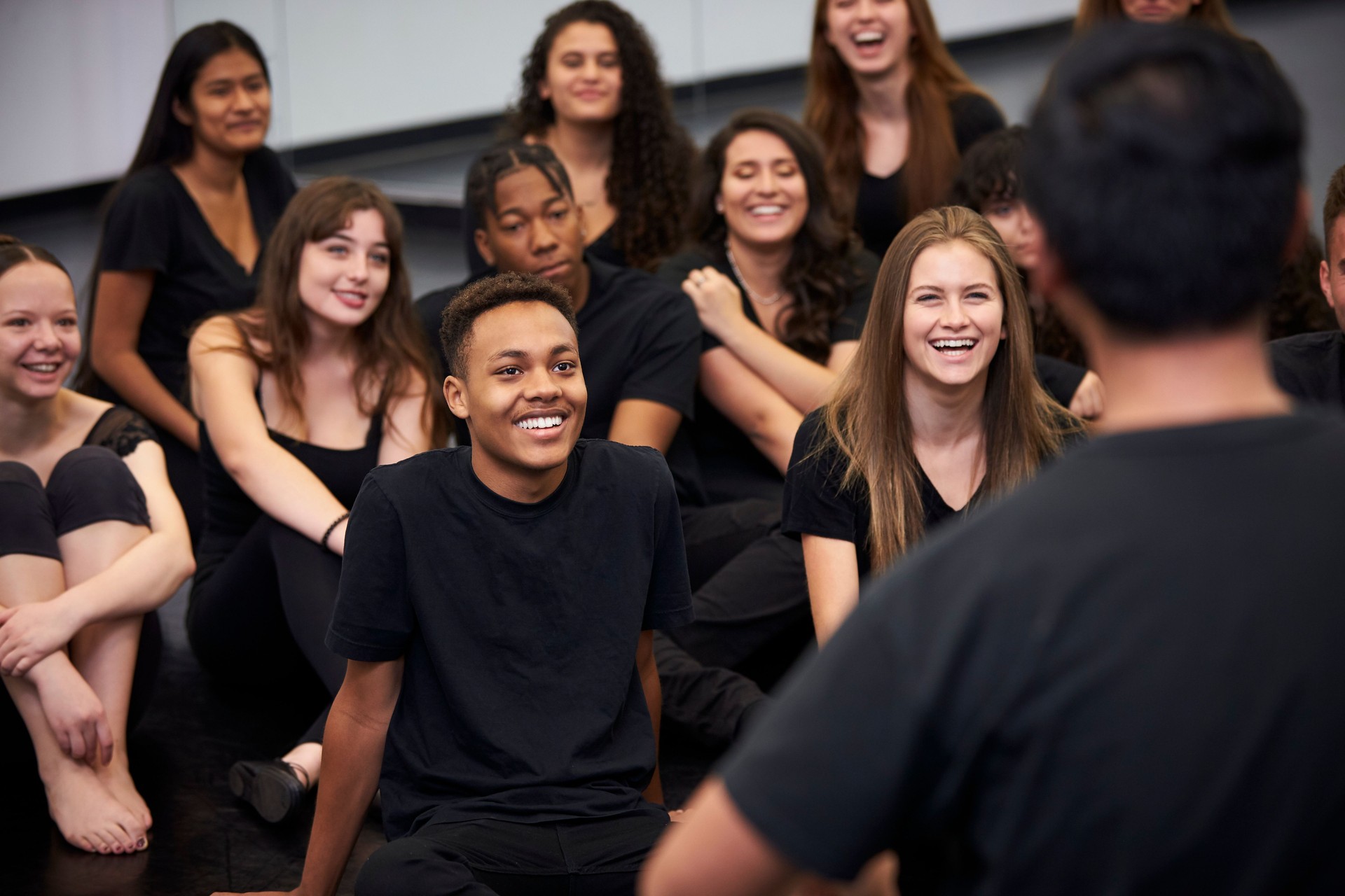 Teacher At Performing Arts School Talking To Students Sitting On Floor In Rehearsal Studio